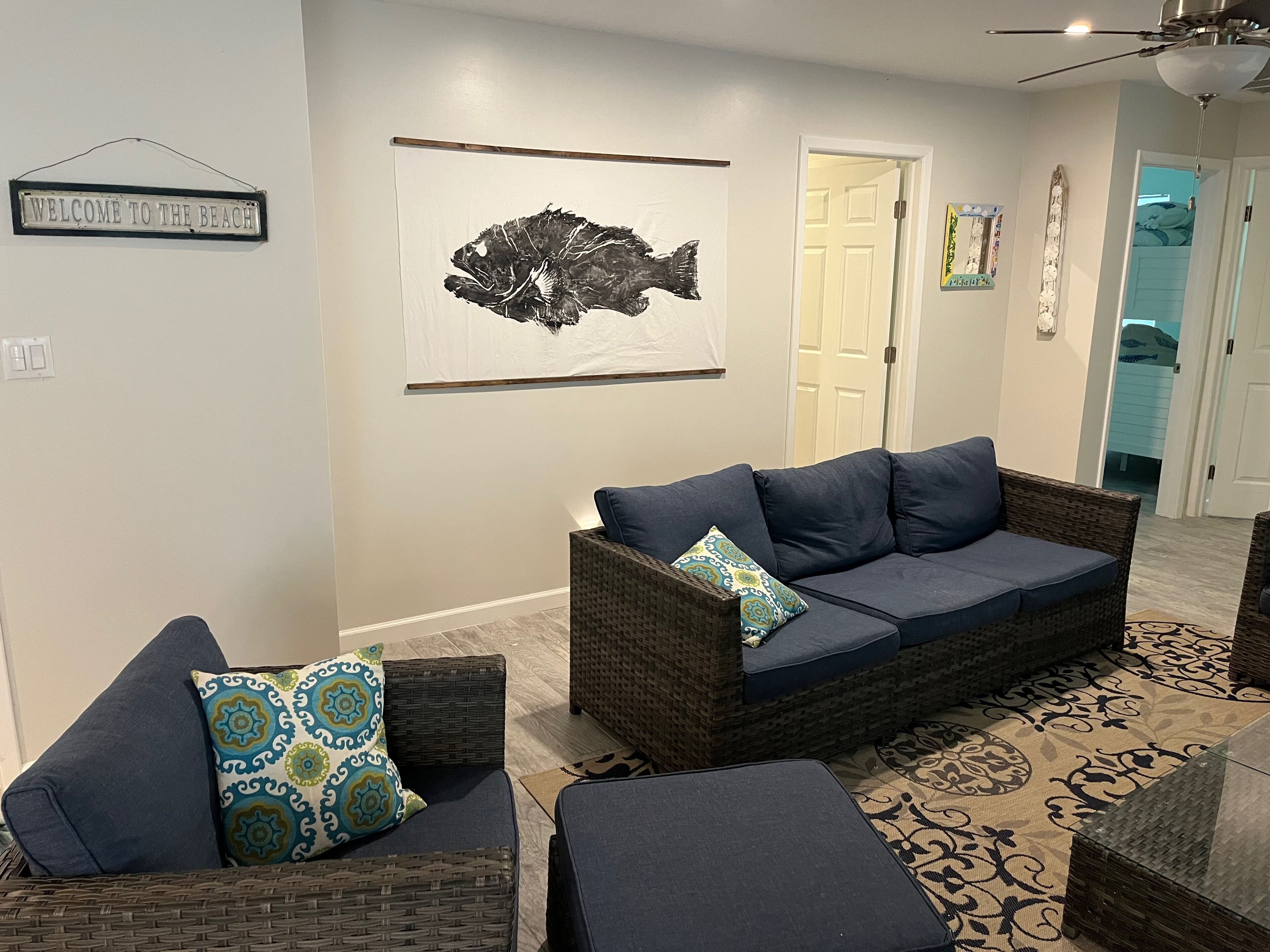 A living room with a dark blue sofa, two matching armchairs, and a patterned rug. A large fish artwork hangs on the wall, and a 'Welcome to the Beach' sign is visible.