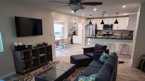 A modern living room with a sofa, coffee table, and entertainment unit. A kitchen and dining area are visible in the background. - The Green Grouper, oversized beach villa. (Central Abaco , Treasure Cay)