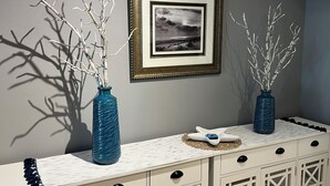 Ein weißer Kommode mit zwei blauen Vasen, die Zweige enthalten, einem gerahmten Foto an der Wand und einem kleinen dekorativen Gegenstand auf der Kommode.