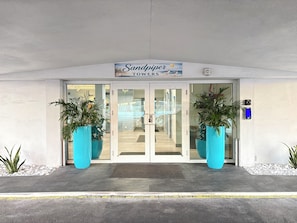 A hotel entrance with a sign reading 'Sandpiper Towers,' featuring a glass door, two large potted plants, and a metal security device on the wall.