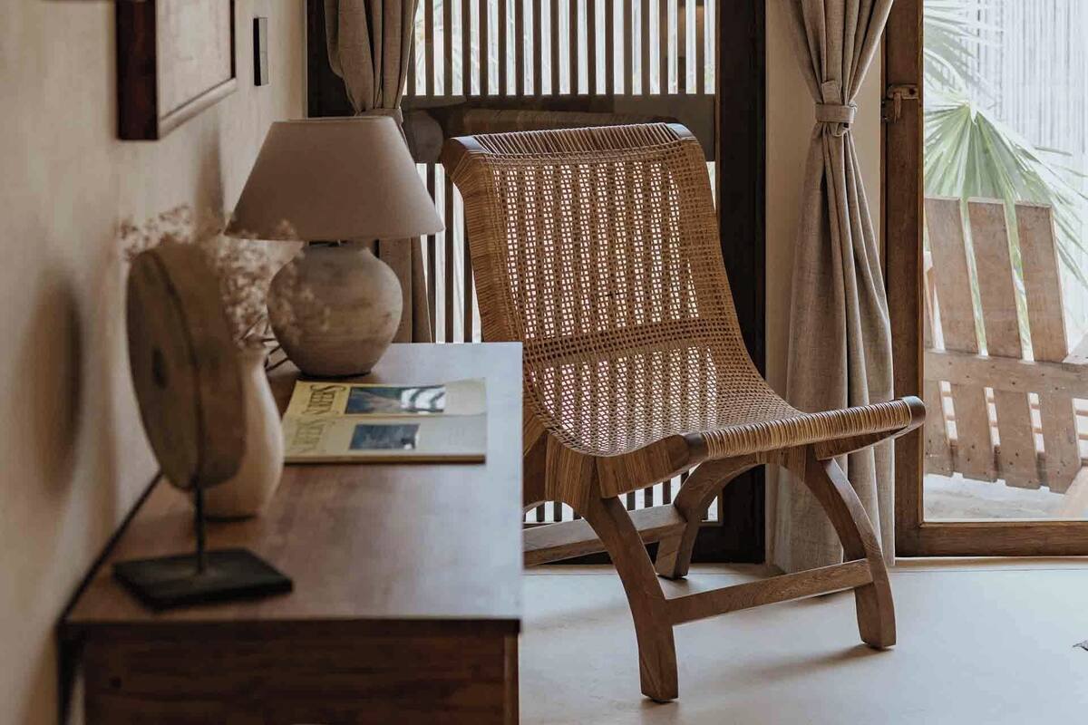 Une pièce avec une chaise en bois, une table avec une lampe et une fenêtre avec des rideaux.