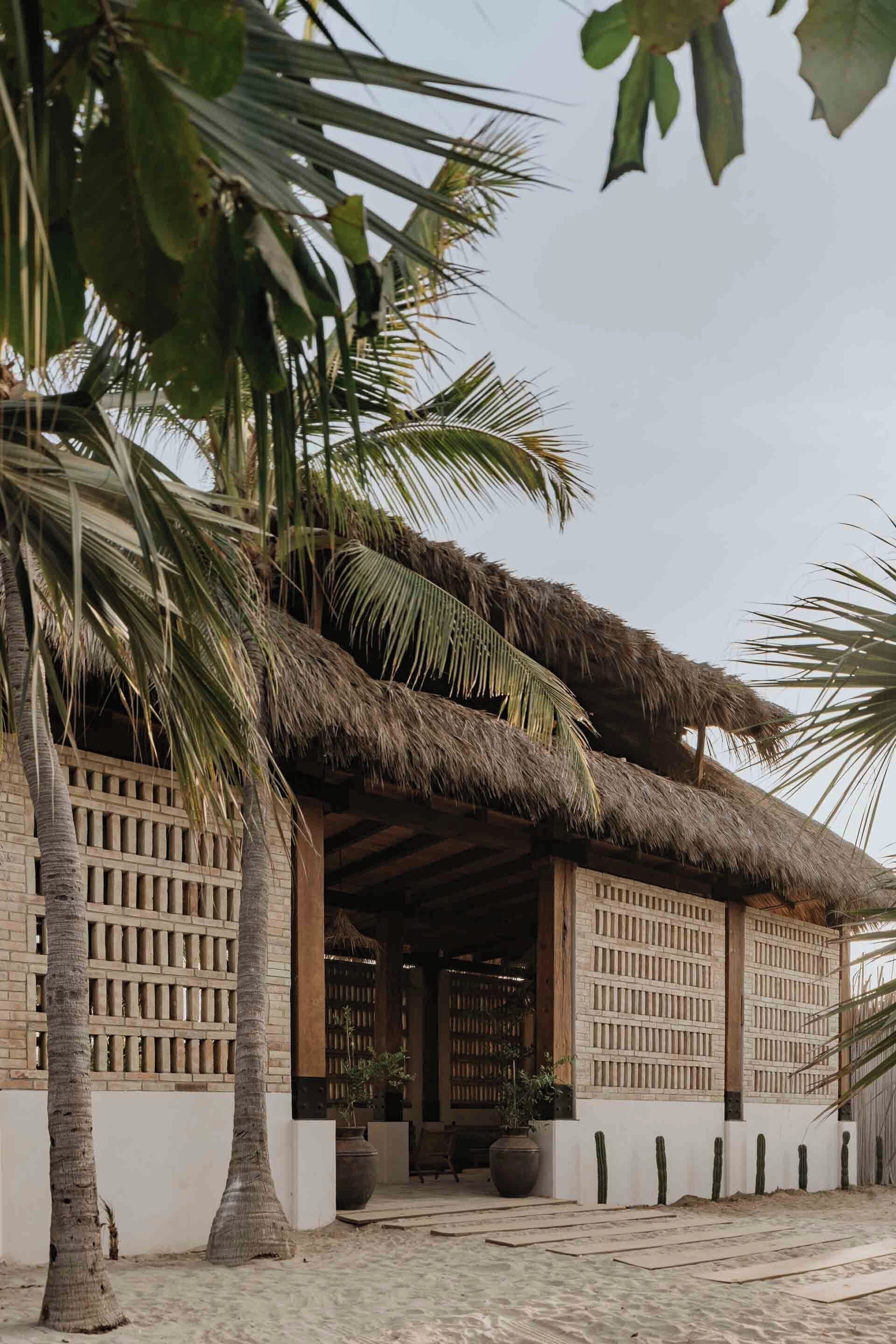 A traditional thatched-roof structure with a wooden lattice wall, surrounded by palm trees and potted plants.