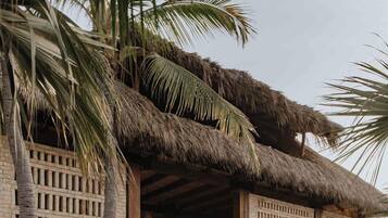 A traditional thatched-roof structure with a wooden lattice wall, surrounded by palm trees and potted plants.