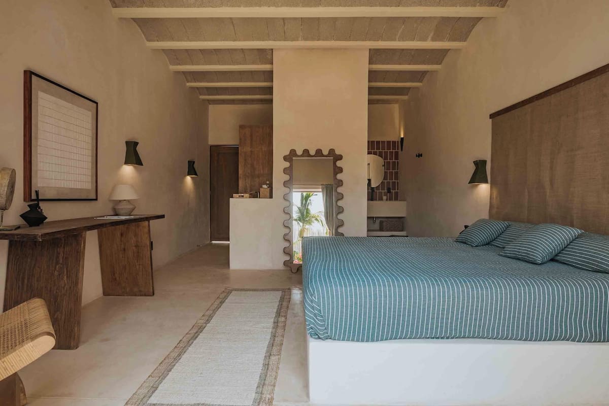 Une chambre avec un grand lit, un bureau et une chaise. Il y a un miroir sur le mur et un tableau encadré au-dessus du bureau.