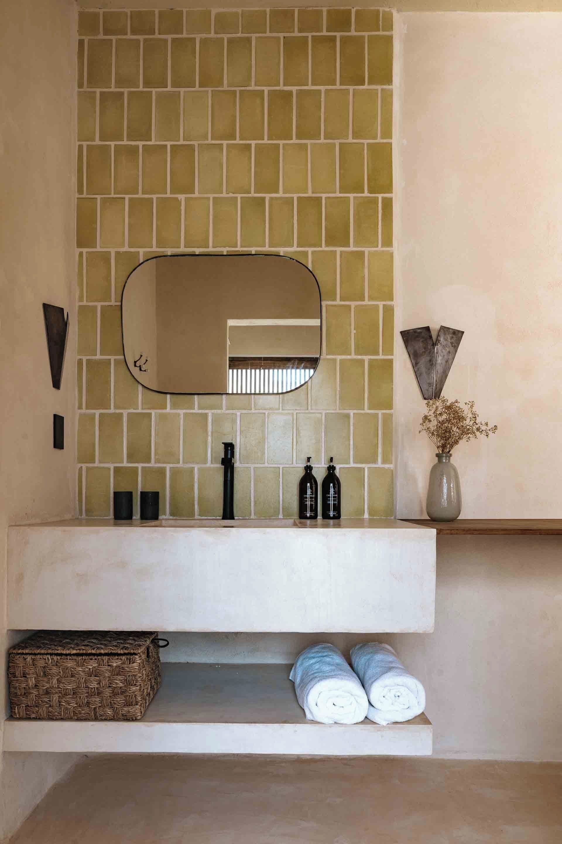 A bathroom with a tiled backsplash, a mirror, a sink, two bottles, a vase with dried flowers, and two rolled towels on a shelf.