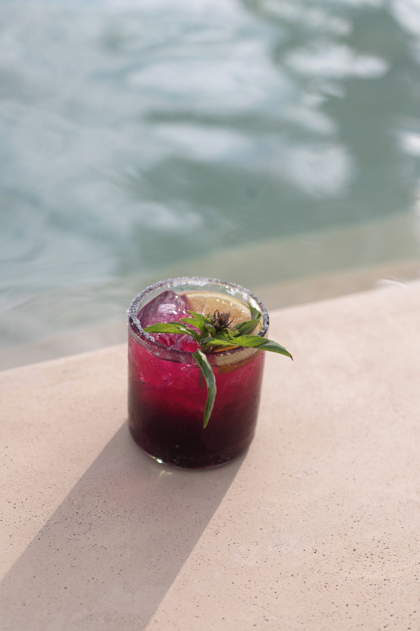 Un vaso de bebida roja, adornado con una hoja de menta, sobre una superficie de hormigón cerca de un cuerpo de agua.