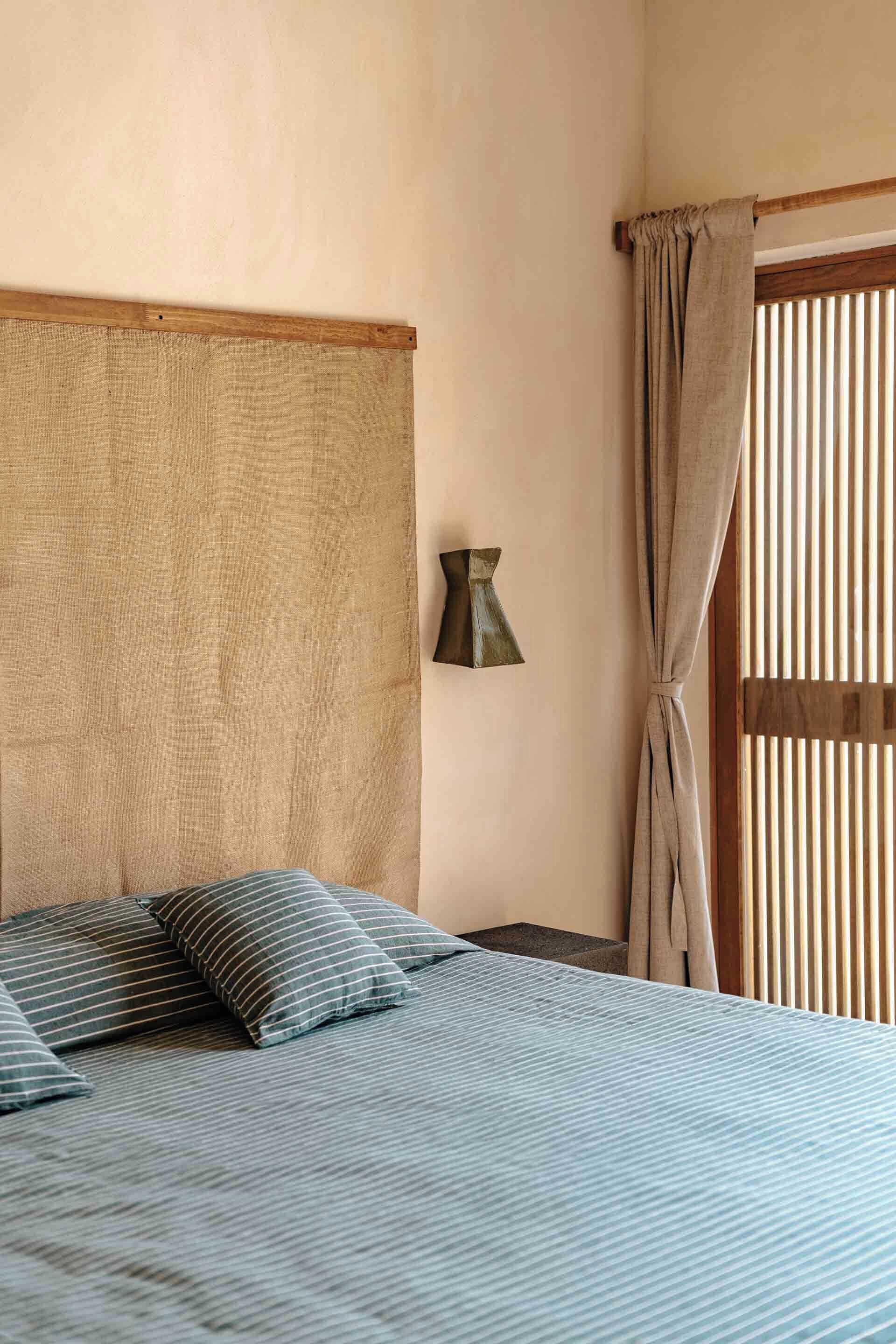 A bedroom with a bed, pillows, a headboard, a wall-mounted lamp, and a window with curtains.