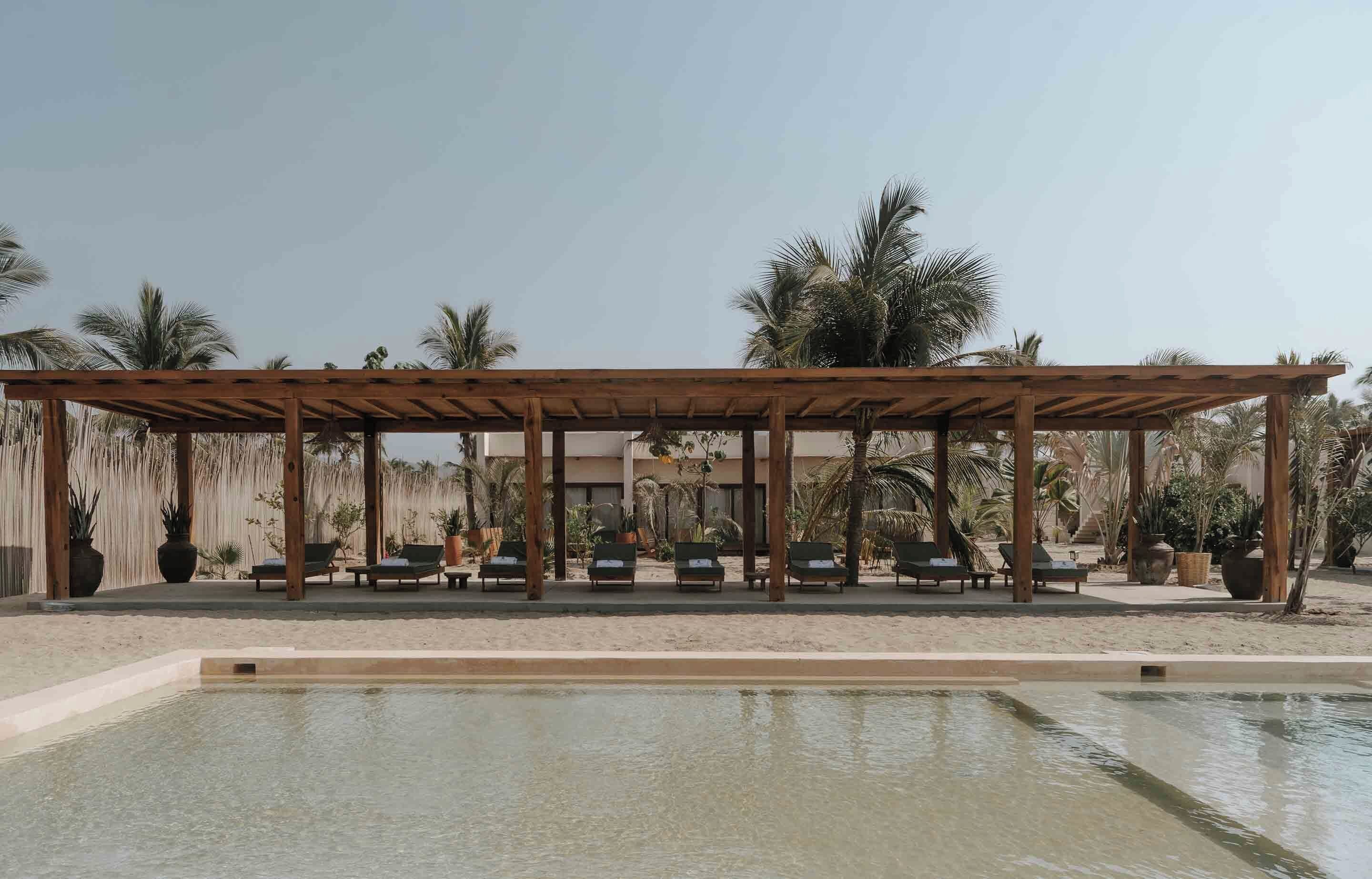A pool area with a wooden pergola, lounge chairs, and palm trees.