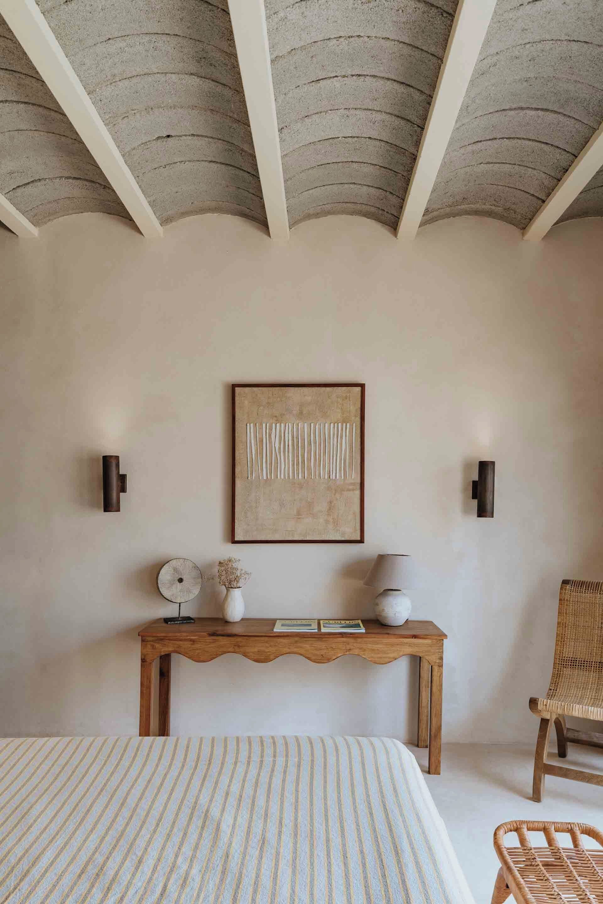 A bedroom with a bed, a wooden console table with decorative items, a wicker chair, and a ceiling with exposed beams.