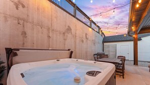 Un bain à remous installé dans un espace patio extérieur, avec un mur en béton, une terrasse en bois et des guirlandes lumineuses.