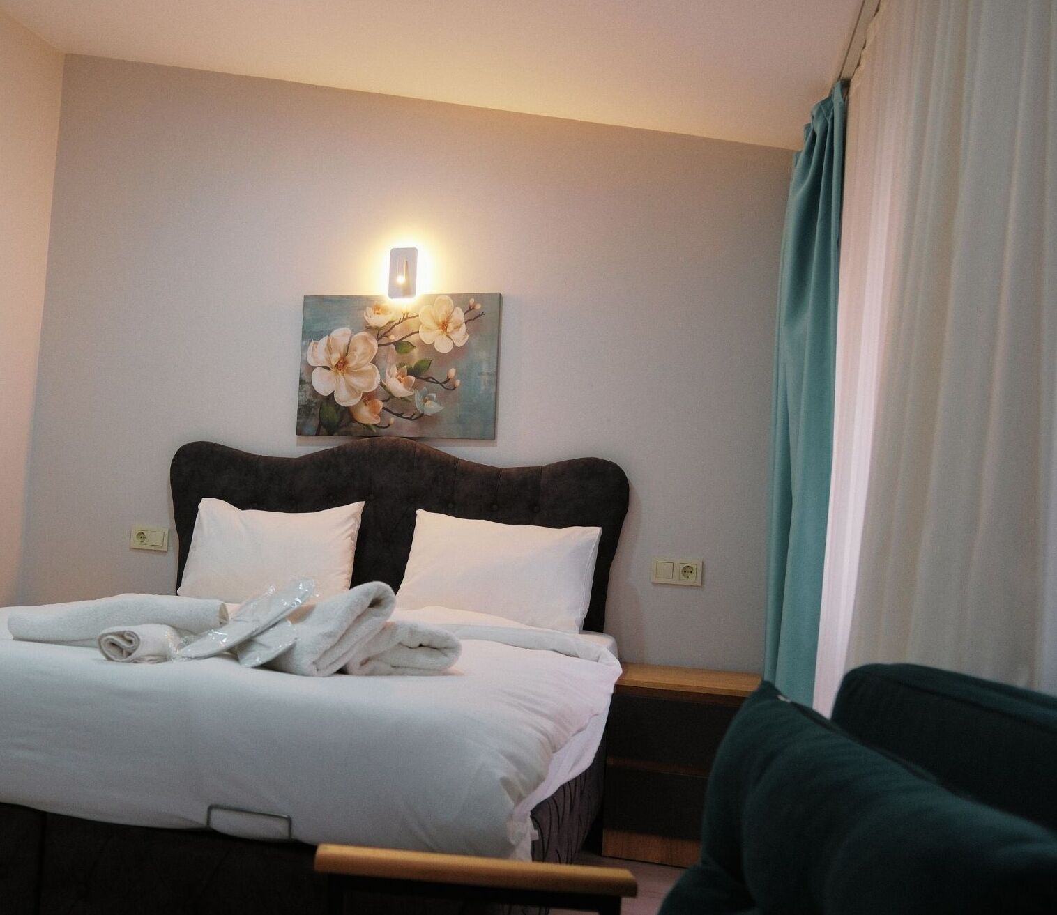 A hotel room with a bed, a painting of flowers, a nightstand, and a green armchair.