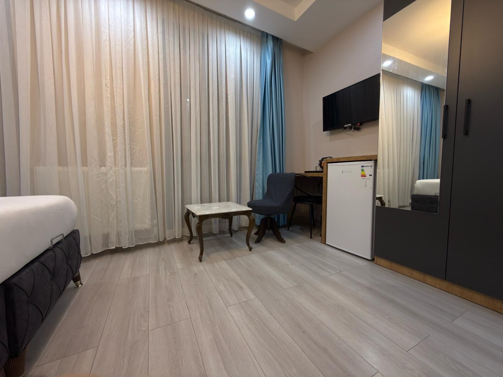 A hotel room with a bed, a small table, a chair, a desk, a refrigerator, and a wardrobe.