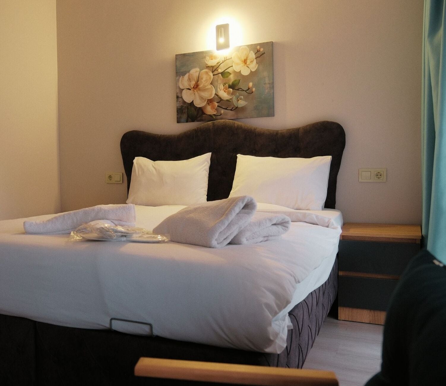 A neatly made bed with white linens and towels, a dark headboard, and a painting of flowers on the wall above it.