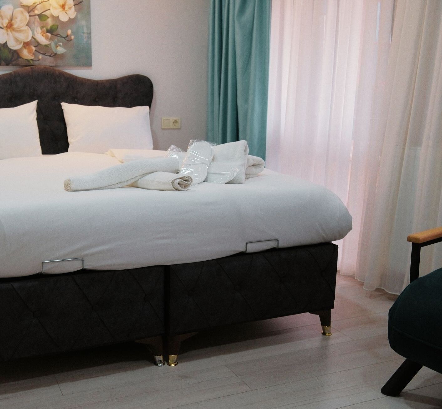 Una cama bien tendida con sábanas blancas y toallas enrolladas, un cabecero oscuro y una cortina color verde azulado al fondo.