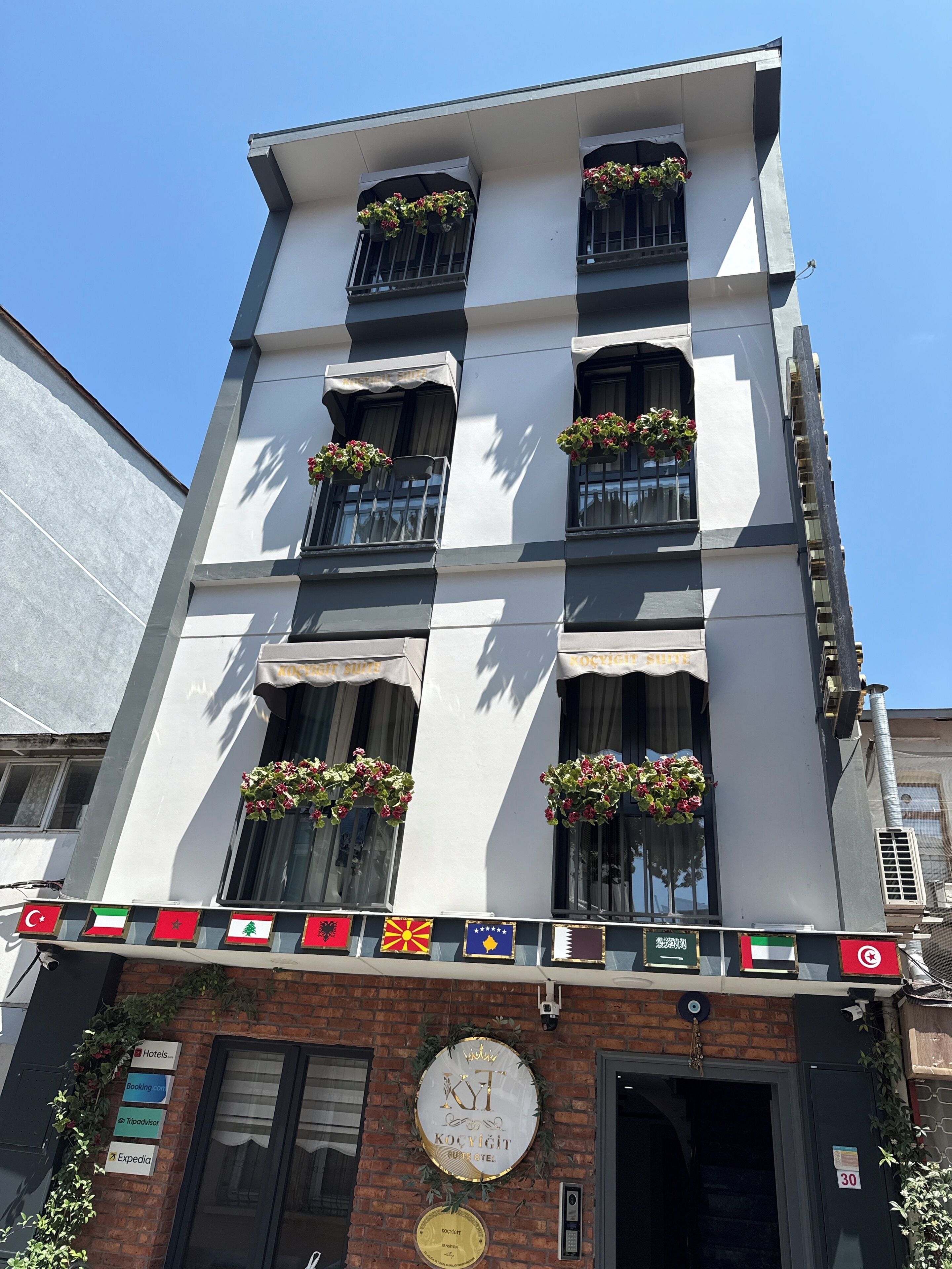 A multi-story hotel with balconies adorned with flower baskets, a signboard with flags, and a door with a round plaque.