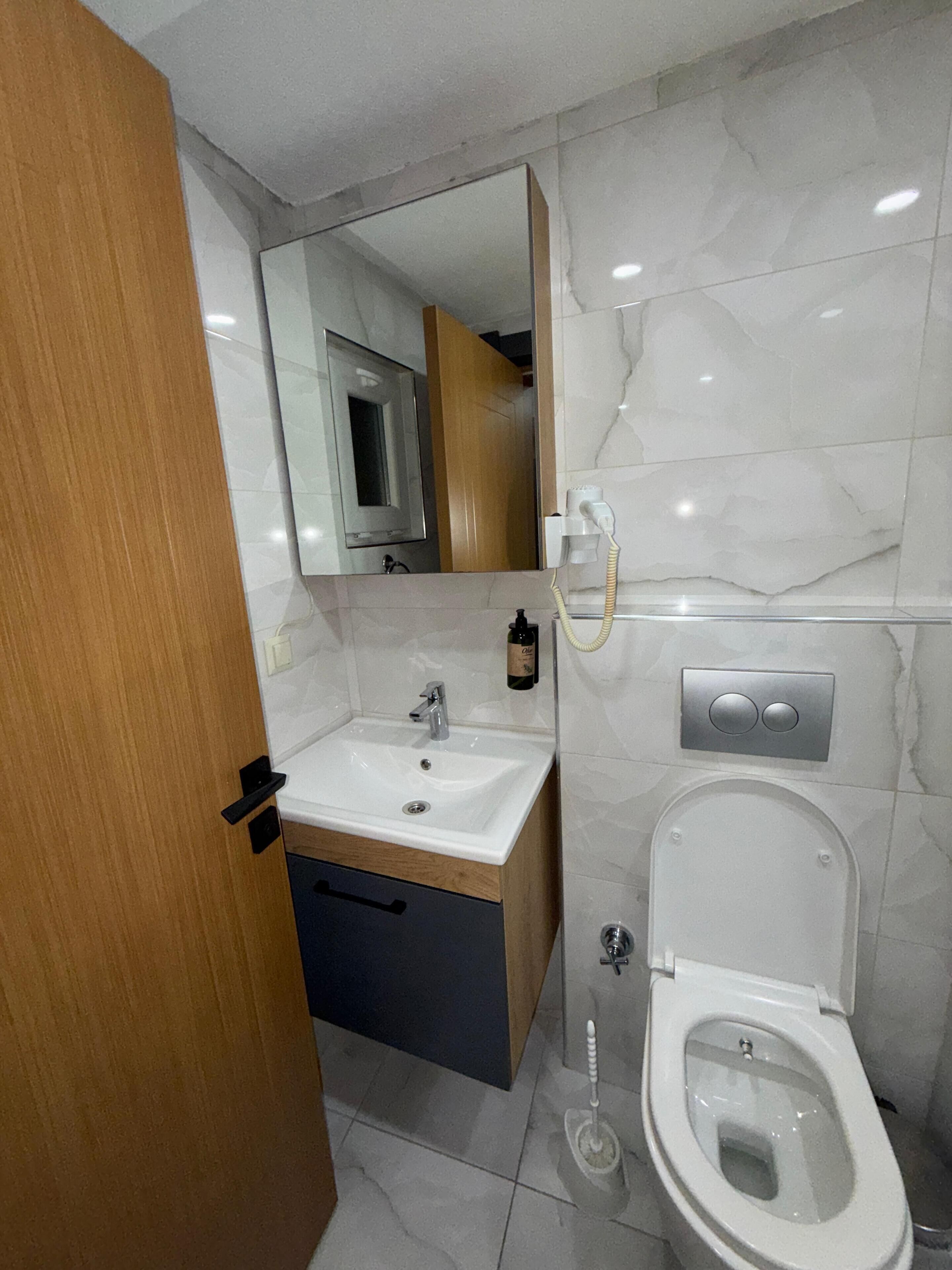 A modern bathroom with a wooden vanity, a white sink, a mirror, a toilet, and a marble wall.