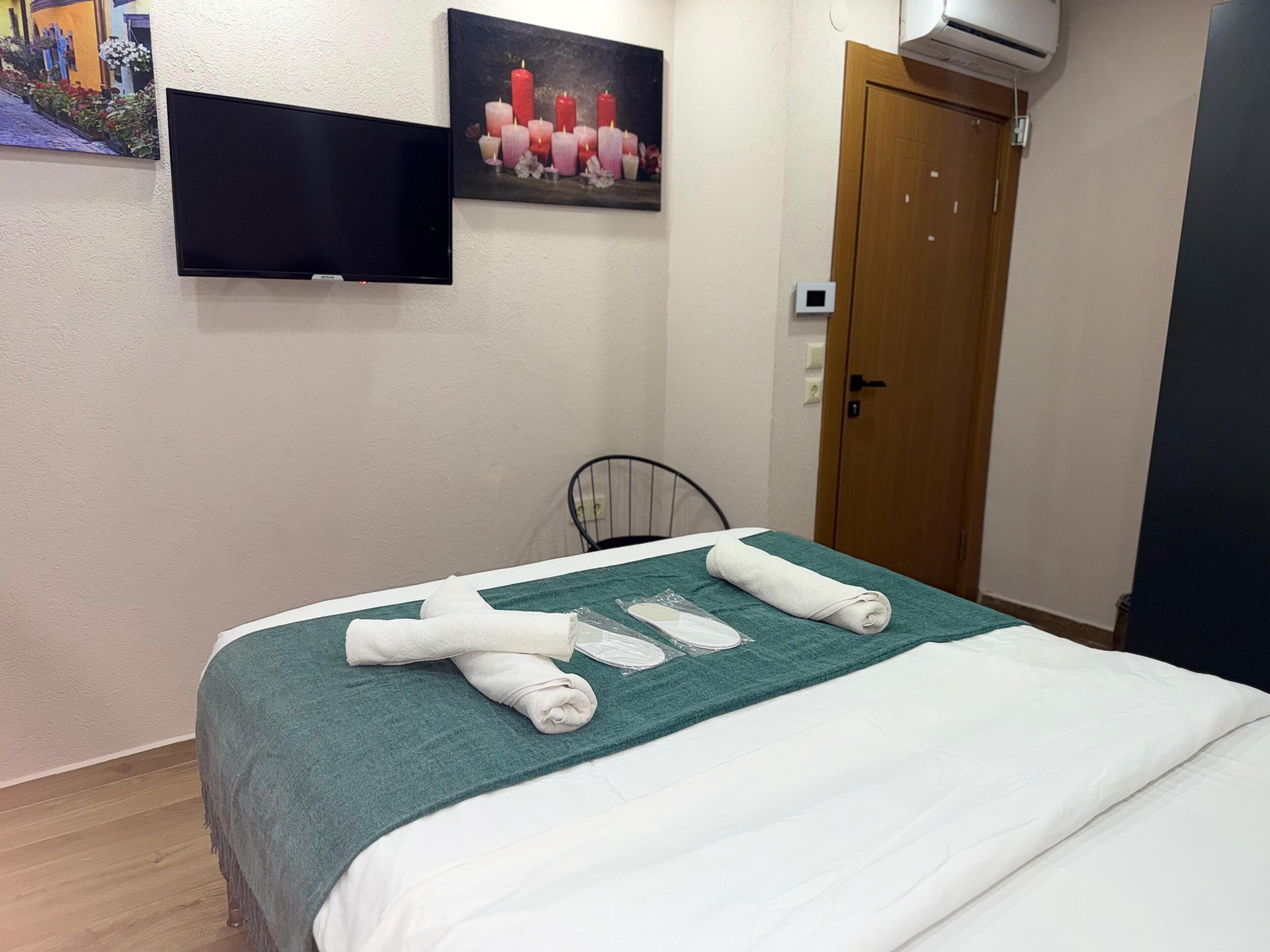 A neatly made bed with a green blanket and white sheets, a flat-screen TV mounted on the wall, and a painting of candles.