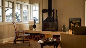 Una habitación acogedora con una chimenea grande, un banco de madera, un sillón de mimbre y vistas al exterior a través de amplias ventanas.