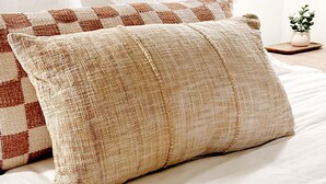 Un llit amb roba de llit blanca i dos coixins amb textura, una làmpada de paret i una petita planta sobre una tauleta de nit.
