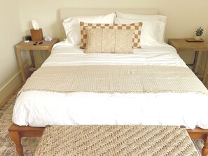 A neatly made bed with a checkered throw pillow, a bedside table with a tissue box and a clock, and a woven bench at the foot of the bed.
