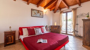 A bedroom with a large bed, wooden ceiling beams, a painting on the wall, and a view through a glass door. - Sea view by Interhome (Muravera Villaputzu)