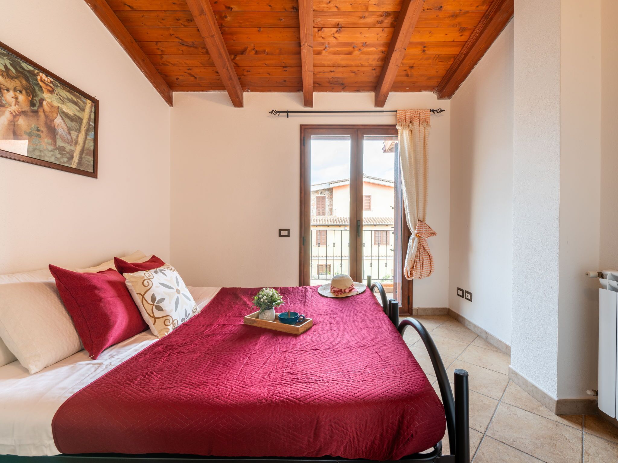 A bedroom with a wooden ceiling, a bed with a red bedspread, a window with curtains, and a view of buildings outside.