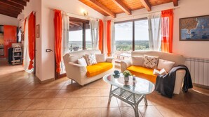 A spacious living room with a high wooden ceiling, two white sofas with orange cushions, a glass coffee table, and a view of the countryside through large windows. - Sea view by Interhome (Muravera Villaputzu)