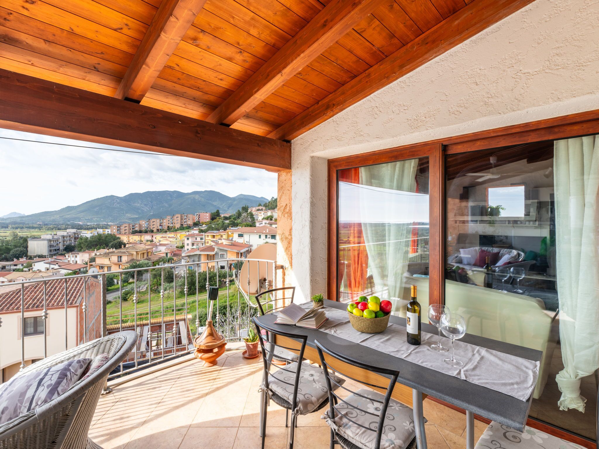 Un balcón con una mesa lista para comer, una silla de mimbre y vistas a un pueblo y montañas.