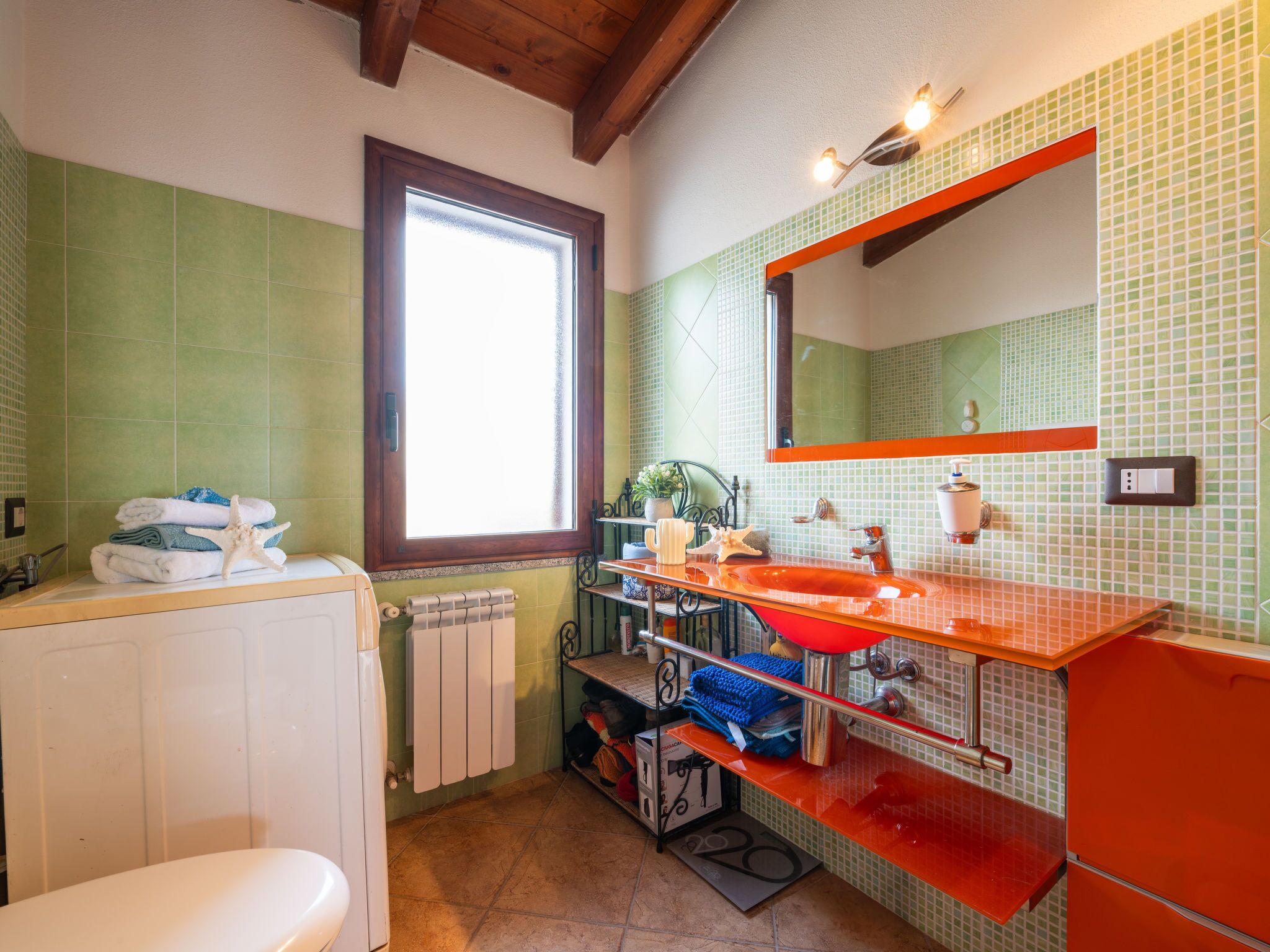 Une salle de bain avec des toilettes blanches, un lavabo rouge et un miroir au-dessus. Il y a une fenêtre donnant sur l’extérieur.