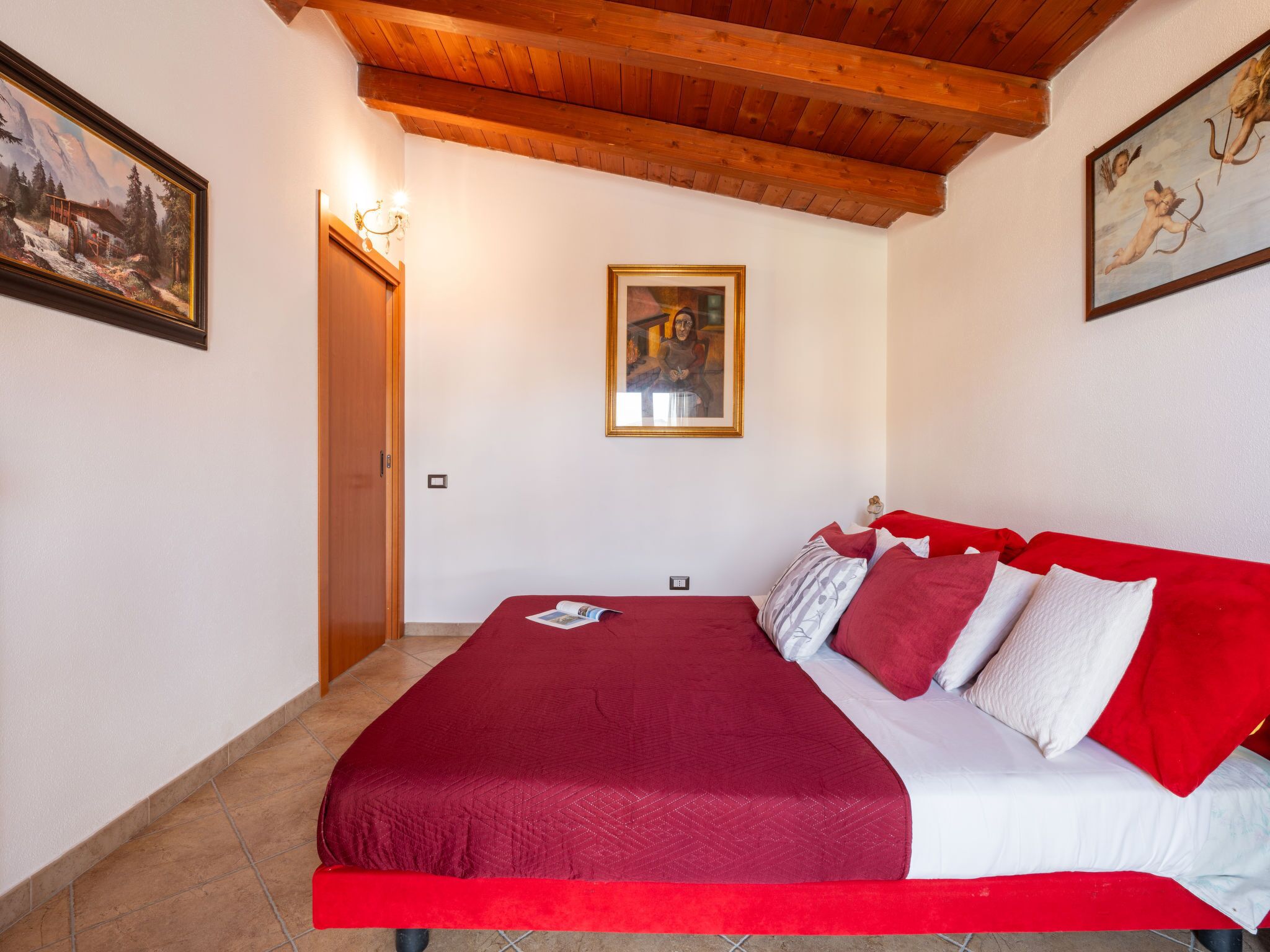A bedroom with a wooden ceiling, a bed with red bedding, and two framed pictures on the walls.