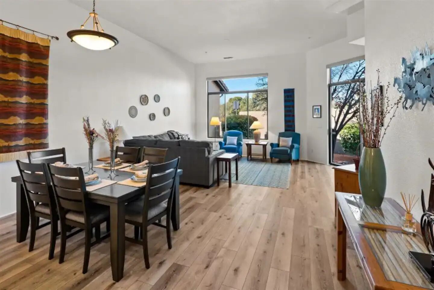 A spacious living area with a dining table, chairs, a sofa, and a coffee table. There is a large window providing natural light and a view of the outdoors.