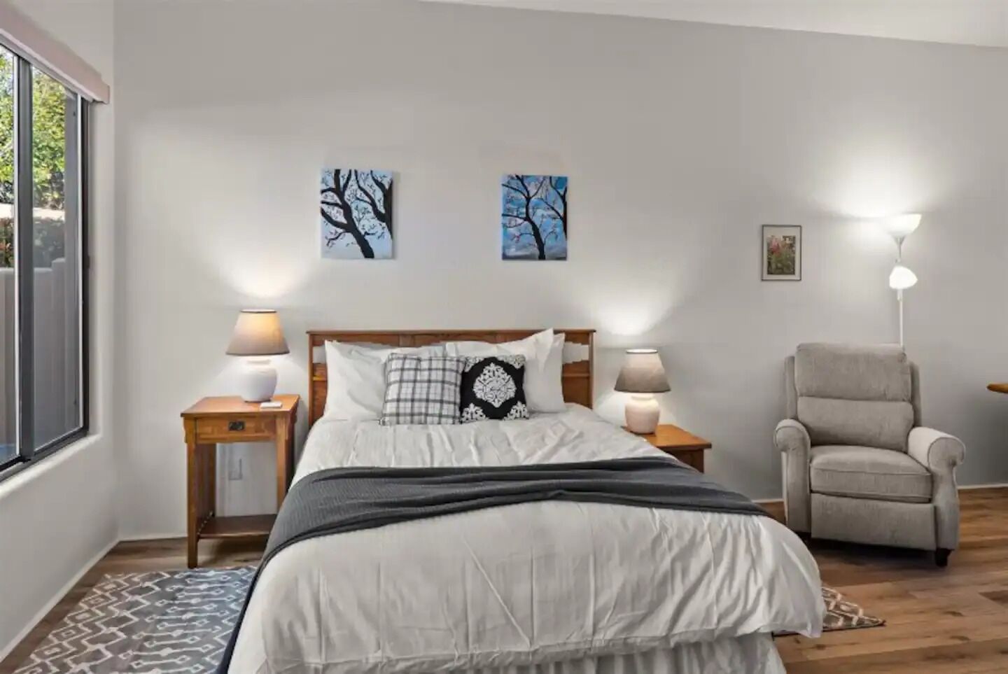 Un dormitorio con una cama, dos mesitas de noche con lámparas, una silla y una obra de arte en la pared.