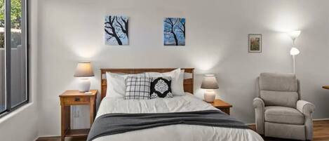 Un dormitorio con una cama, dos mesitas de noche con lámparas, una silla y una obra de arte en la pared.