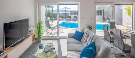 A modern living room with a flat-screen TV, a glass coffee table, a grey sofa with blue cushions, and a view of a swimming pool through large sliding glass doors.