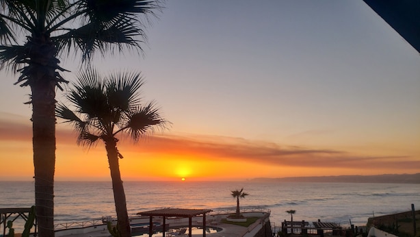 Zonsondergang vanaf een terras met palmbomen, een zwembad en een strand op de achtergrond.