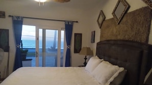 Een slaapkamer met een groot bed, een nachtkastje, een lamp en uitzicht op de oceaan door de gordijnen.