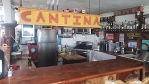 Een barruimte met een 'CANTINA' bord, een koelkast, een fornuis en verschillende flessen en glazen op planken.