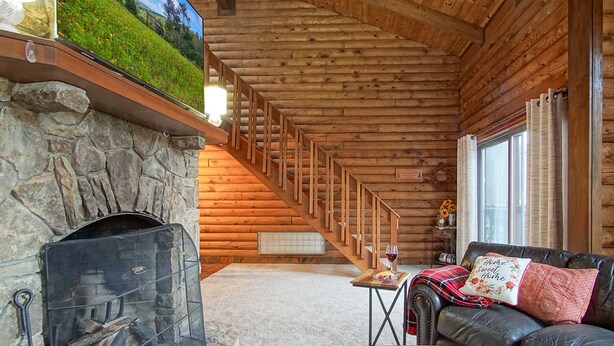 L’intérieur d’une cabane en rondins, avec une cheminée en pierre, un escalier en bois, un canapé en cuir et une vue sur une colline verdoyante par la fenêtre.