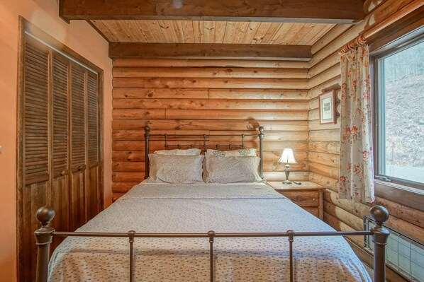 A wooden log cabin bedroom with a bed, a nightstand, a lamp, and a window with curtains. - 2 Br Plus Loft: Hawthorne Valley 4 (Slatyfork)