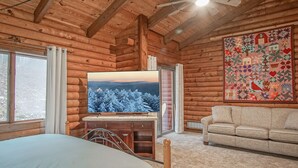 L’intérieur d’une cabane en rondins, avec un lit, un téléviseur posé sur un meuble en bois, un canapé et une grande courtepointe accrochée au mur.