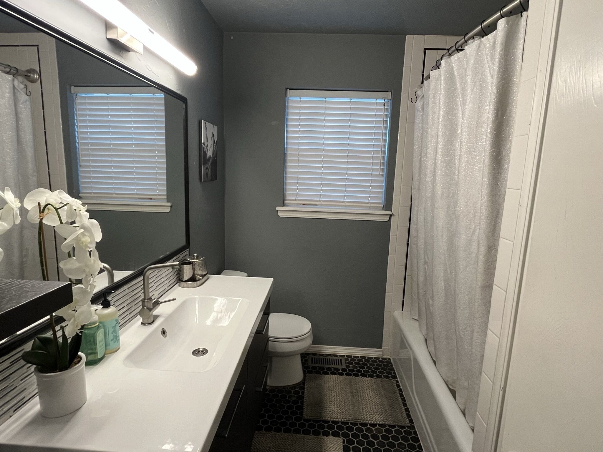 A bathroom with a white sink, a toilet, a shower with a white curtain, and two windows with blinds.
