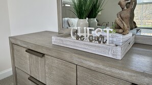 Un aparador de madera con un espejo, una planta y objetos decorativos, incluyendo una figura de oso y un cartel que dice "sean nuestros invitados".