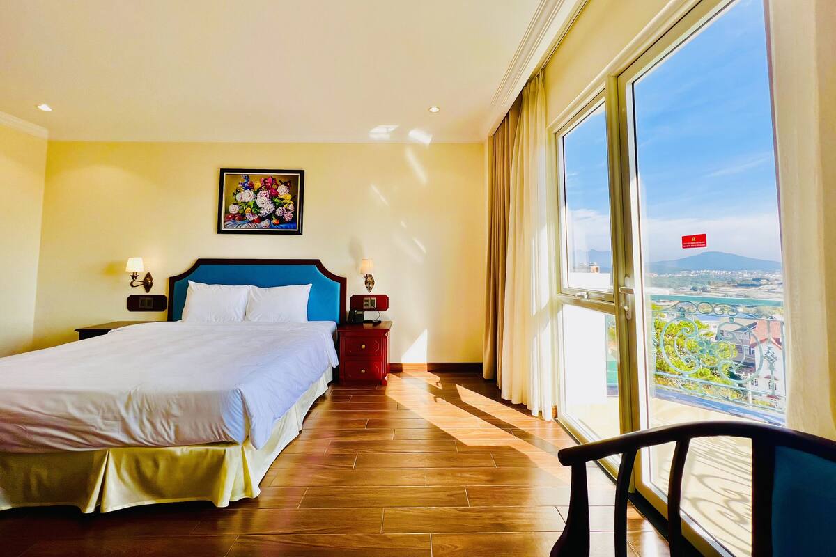 A hotel room with a large bed, a nightstand with a lamp, a painting on the wall, and a balcony with a view.