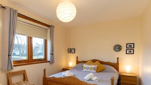 A bedroom with a wooden bed, a bedside table with a lamp, a chair, and a window with curtains. - Culduthel by Interhome (Aviemore)