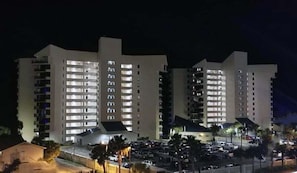 A multi-story hotel with illuminated windows, a parking lot, and a small building with a sloped roof. - BeachHead Paradise-Your Perfect Place in Paradise, Beachfront 1/1! (Panama City Beach)
