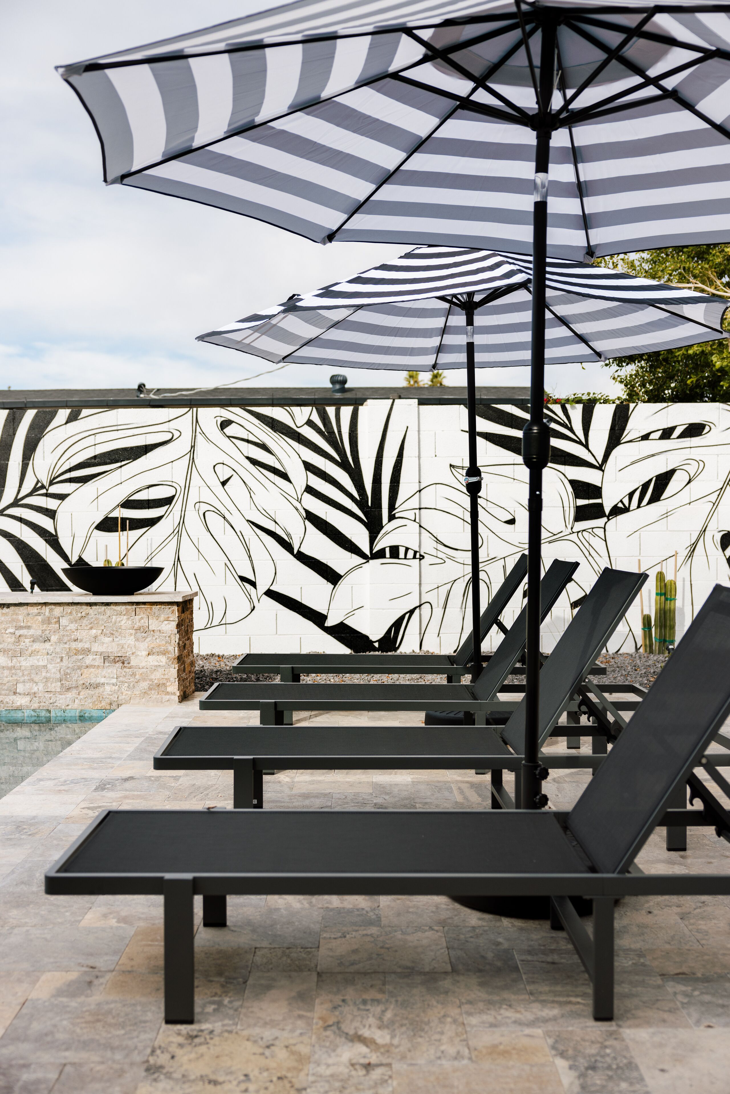 Des fauteuils de jardin, des parasols à rayures, un mur en pierre orné d’une fresque représentant des feuilles et un espace piscine.