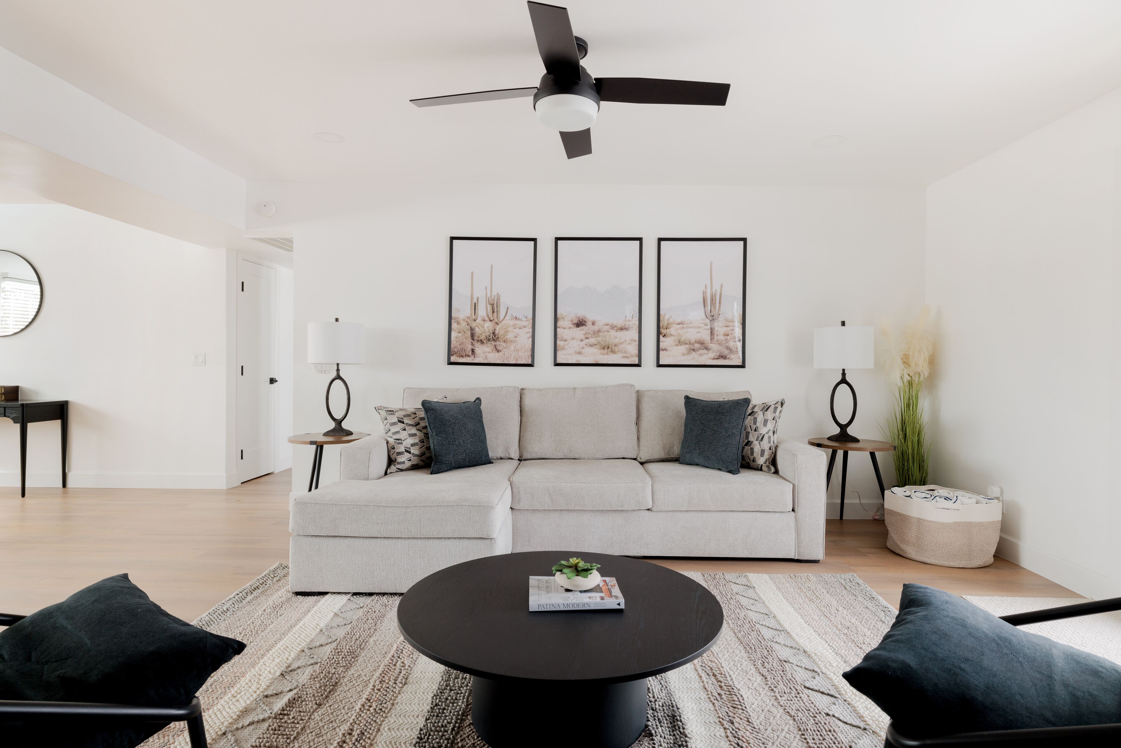 Un salon moderne avec un canapé gris, une table basse noire et un ventilateur de plafond. Trois tableaux encadrés ornent le mur, et une petite table d’appoint avec une lampe est placée à proximité.
