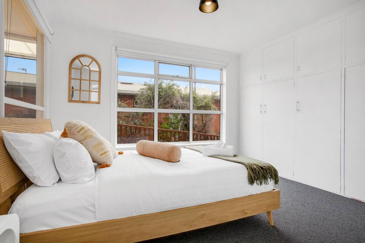 A bedroom with a large bed, white walls, a window with a view of greenery, and a wardrobe.
