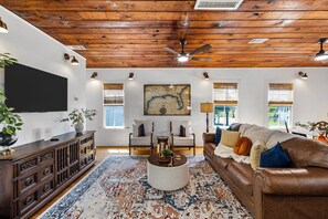 A living room with a wooden ceiling, a large flat-screen TV, a brown leather sofa, a patterned rug, and a map on the wall.