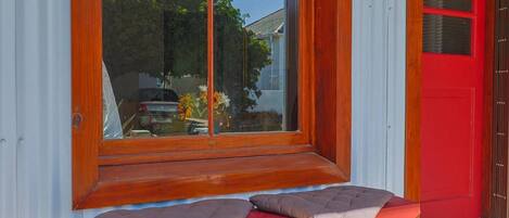 A bench with cushions in front of a window, a red door, and a view of a house and trees.